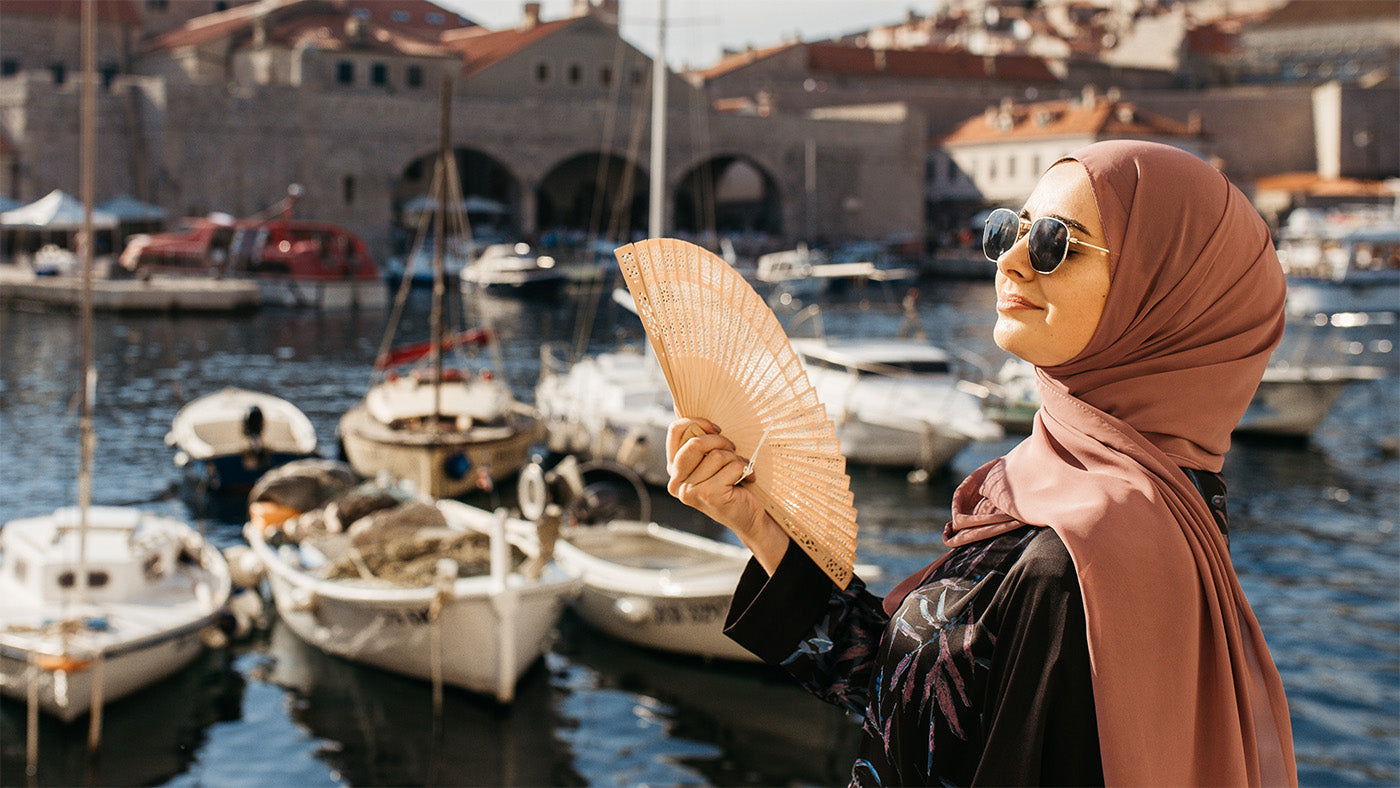 Muslima mit rosafarbenem Hijab und Sonnenbrille am Hafen, schützt sich mit Fächer vor Sommerhitze und trägt Sommer Hijab